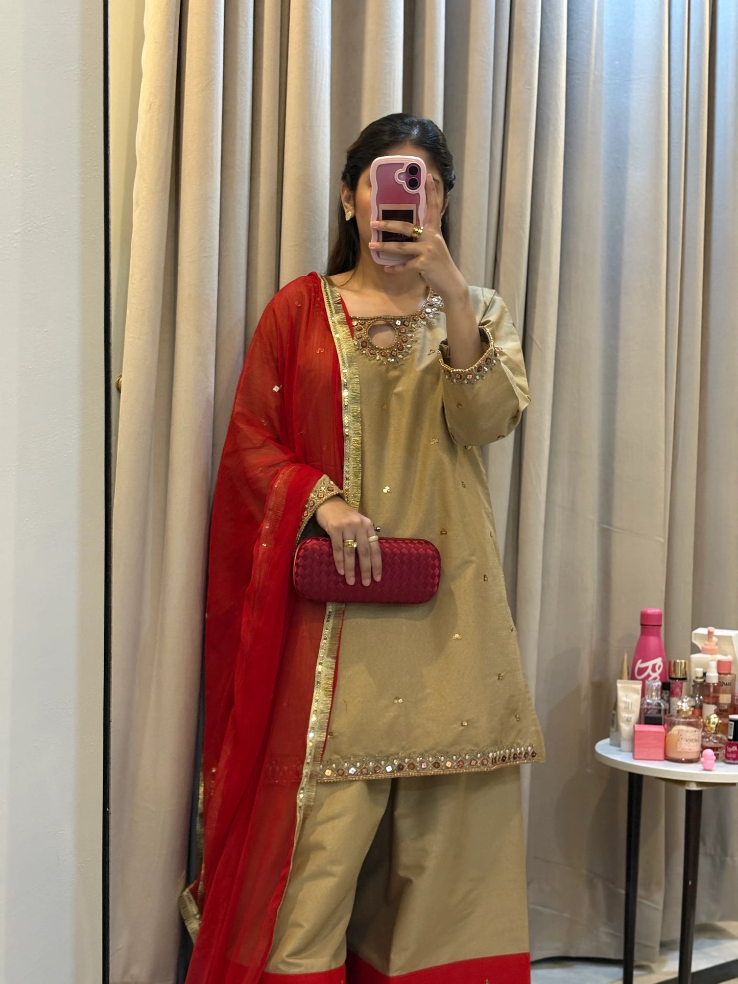 Person wearing a traditional outfit with a red dupatta in front of a curtain.