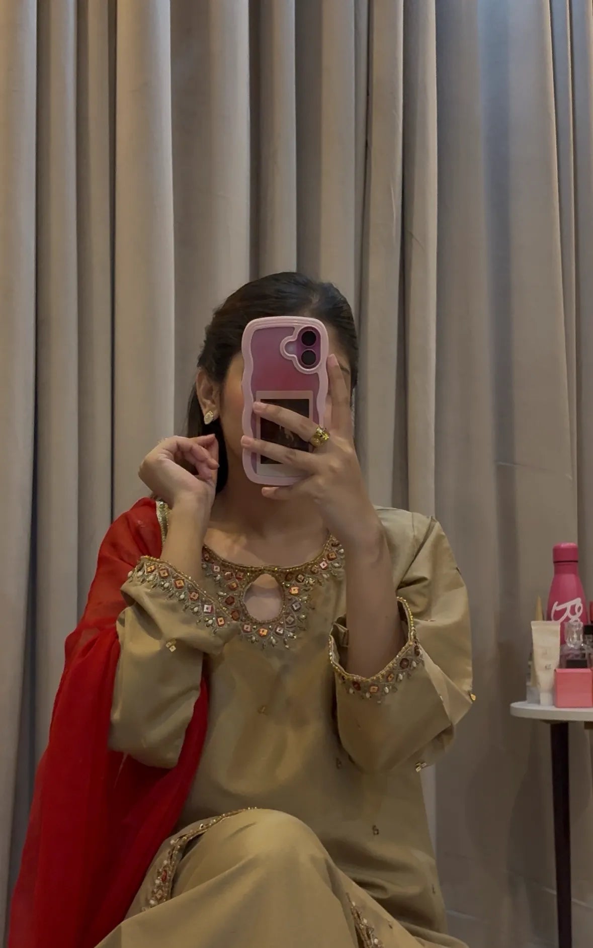 Person wearing a beige traditional outfit with red dupatta sitting against a curtain background.