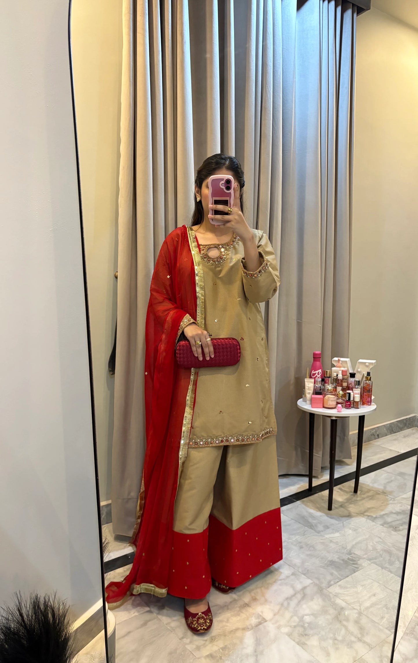 Person wearing a traditional outfit with a red dupatta in a room with a mirror and table.