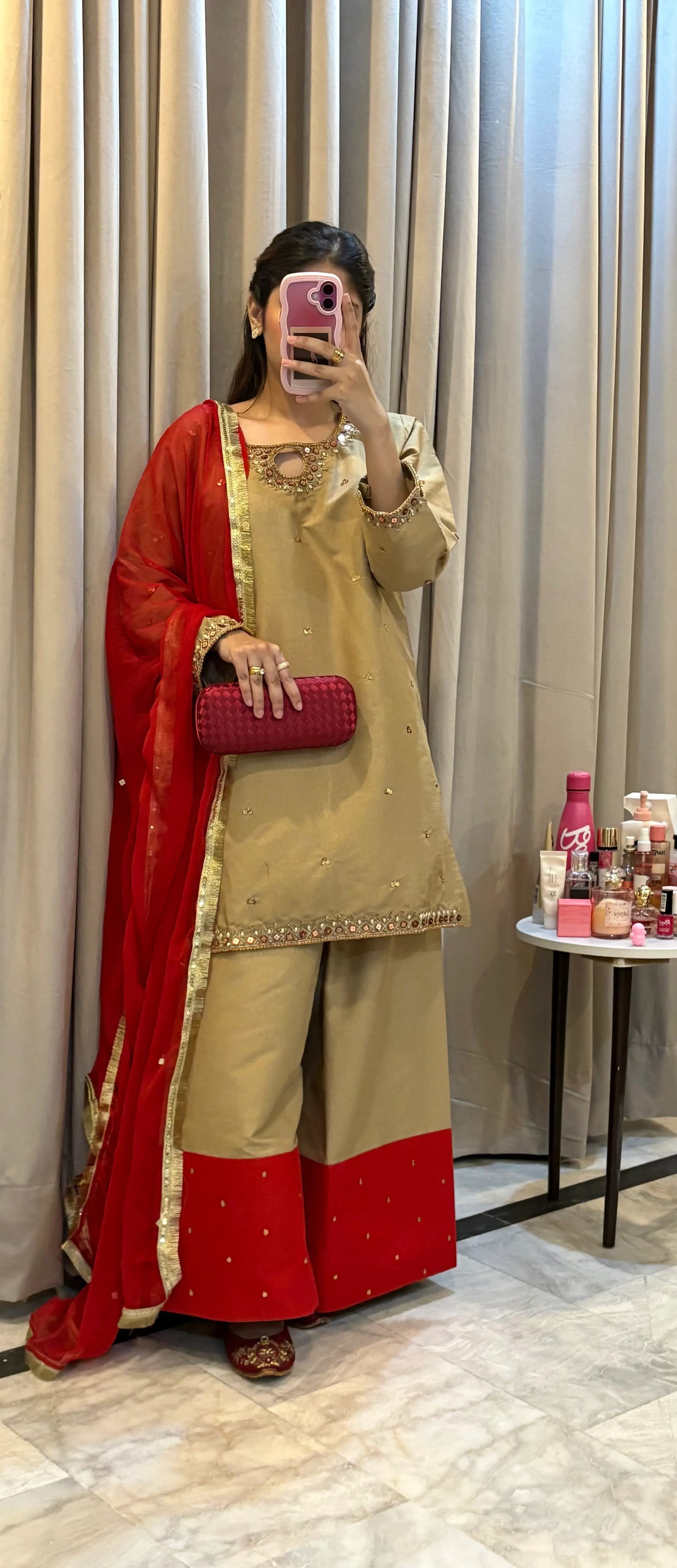 Woman taking a mirror selfie wearing a red and beige traditional outfit in a room with curtains and a small table.