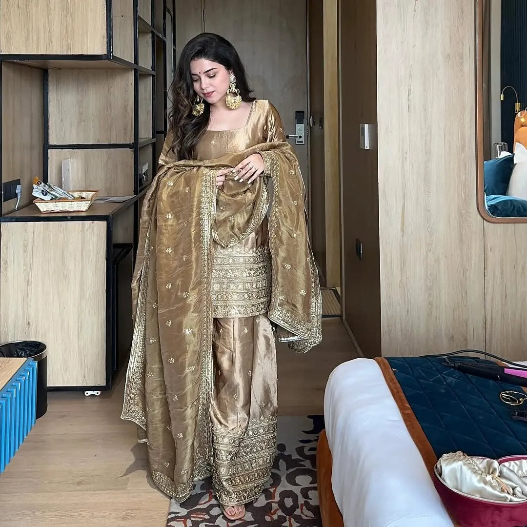Woman in a gold traditional outfit standing in a room with wooden furniture.