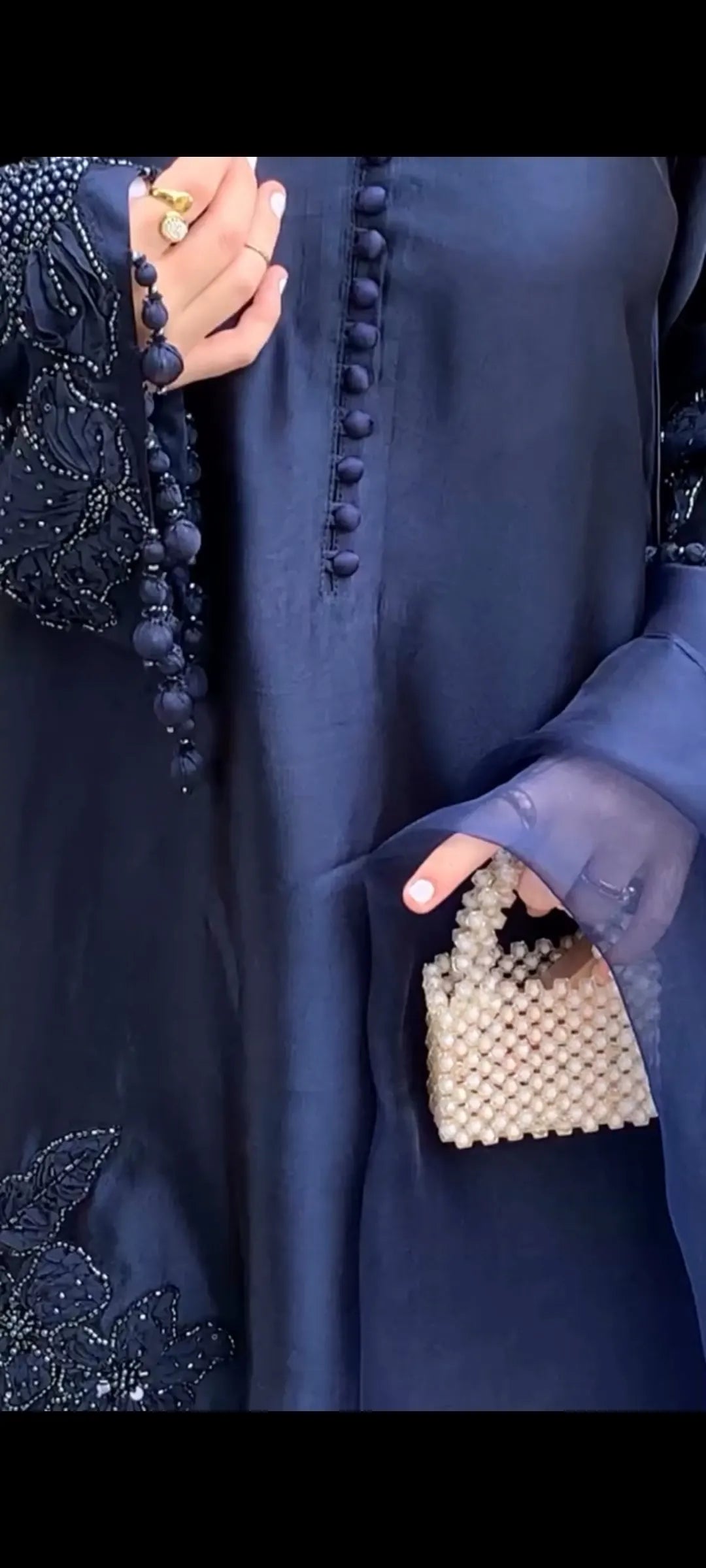Person wearing a dark blue abaya with intricate detailing, holding a beaded handbag.