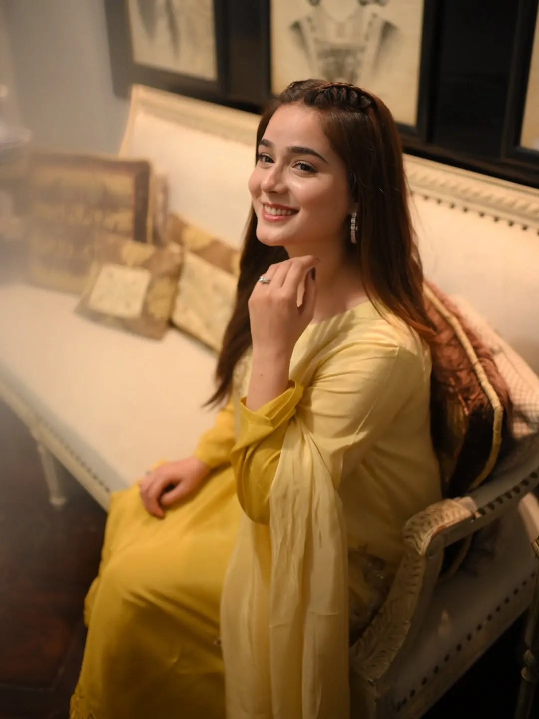 Woman in a yellow dress sitting on a couch in a room with decorative pillows and framed pictures.