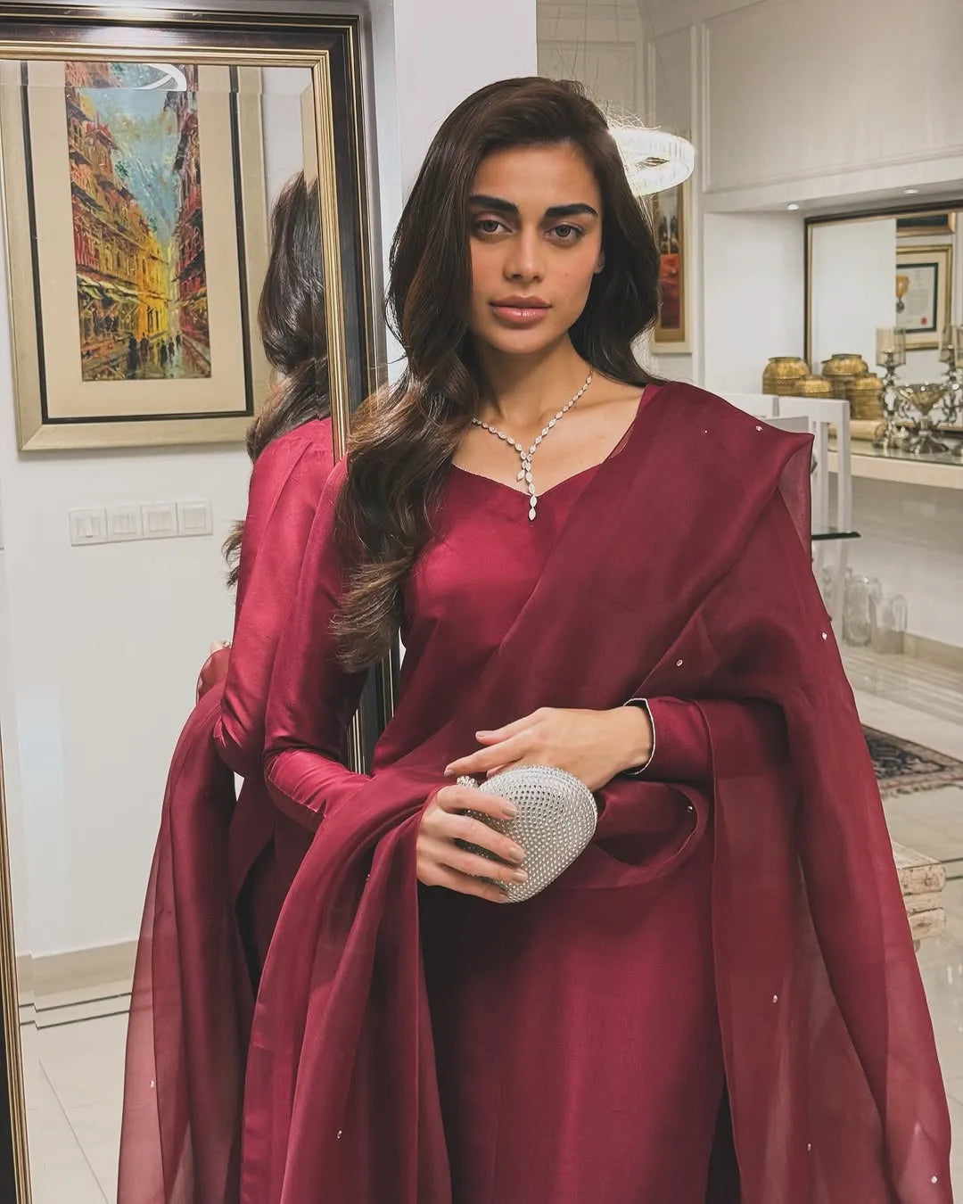 Woman in a maroon saree holding a clutch in a room with decor elements.