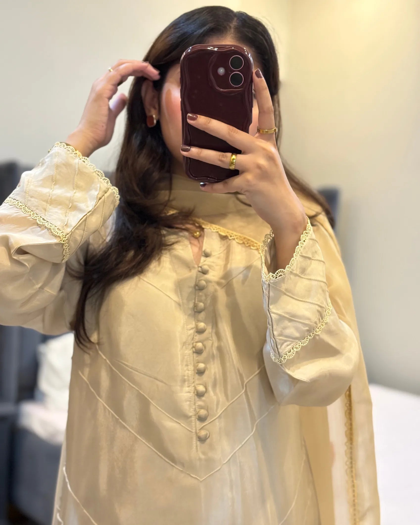 Person wearing a light-colored traditional outfit with intricate designs, holding a phone to take a mirror selfie.