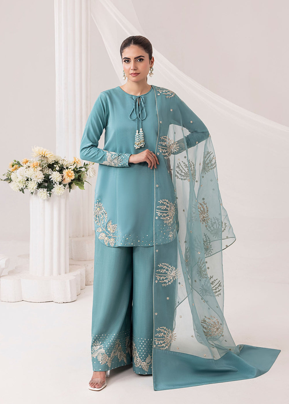 Woman wearing a teal traditional outfit with a matching dupatta in a decorated room.