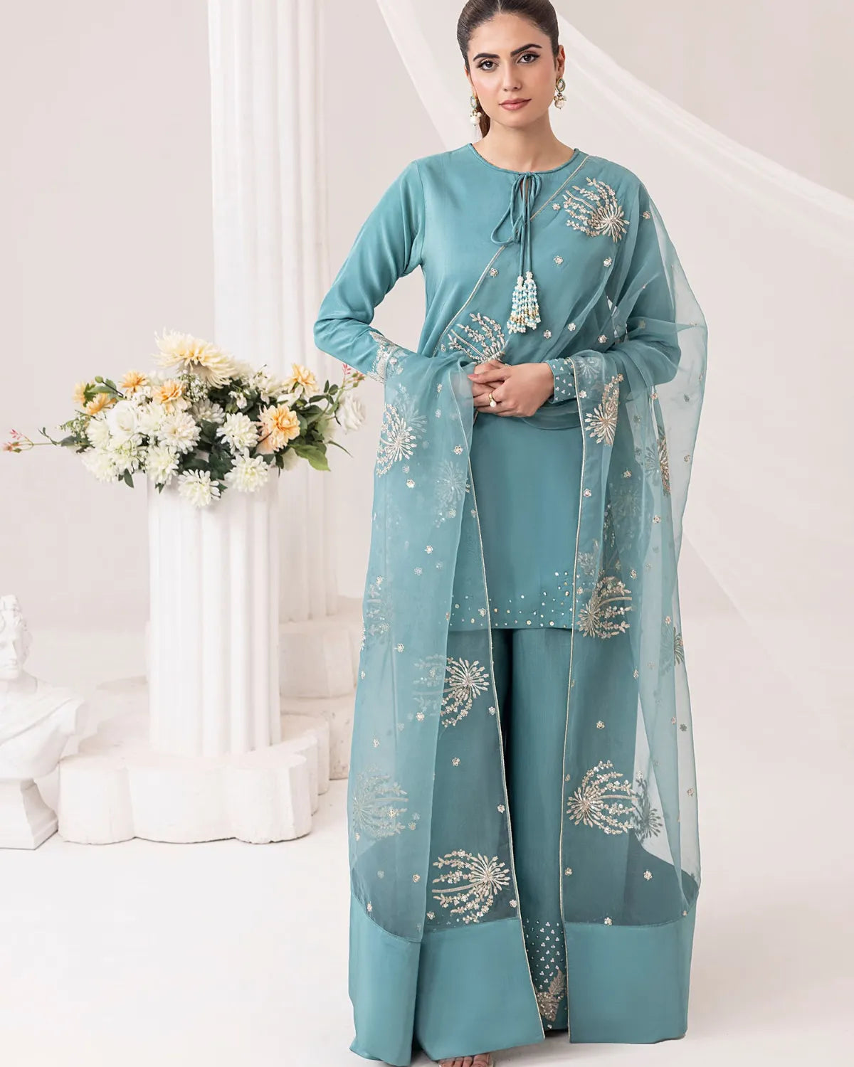 Woman wearing a teal traditional outfit with floral patterns in a white room.