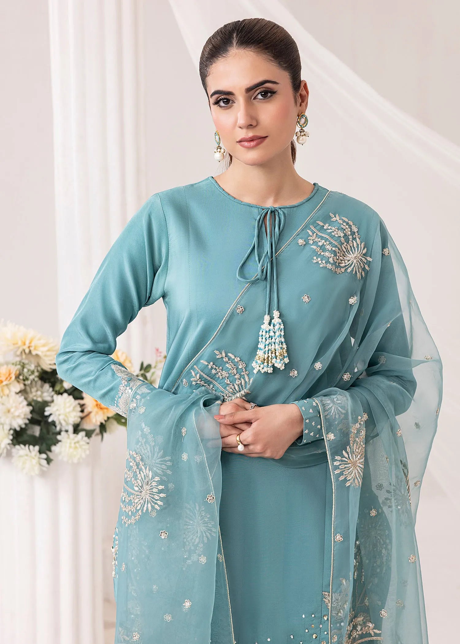 Woman wearing a light blue traditional outfit with floral embroidery, standing indoors.