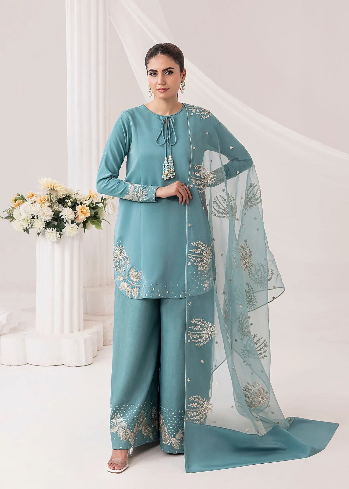 Woman wearing a teal traditional outfit with a matching dupatta in a decorated room.