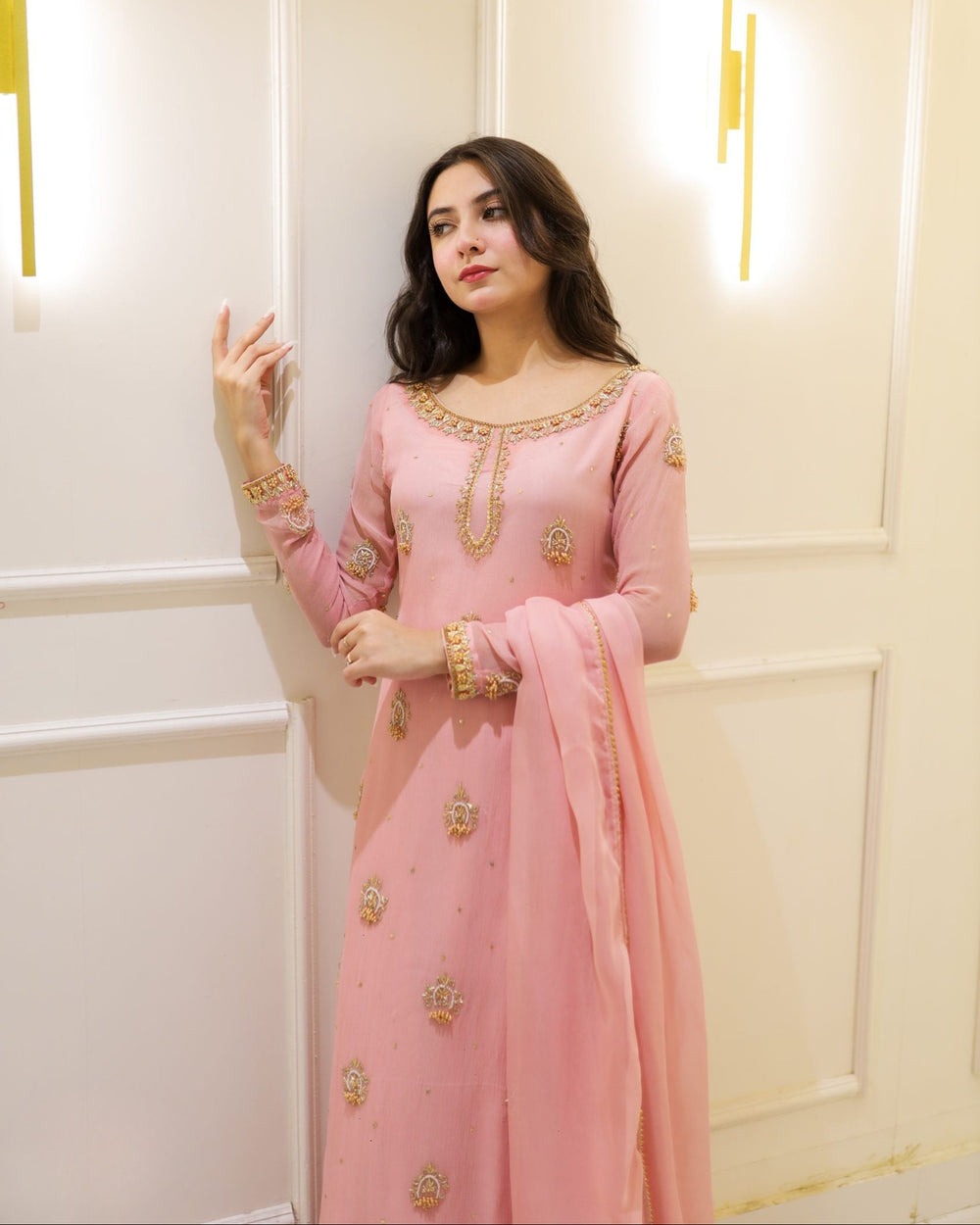 Woman in a pink traditional outfit with gold embroidery standing against a light-colored wall.