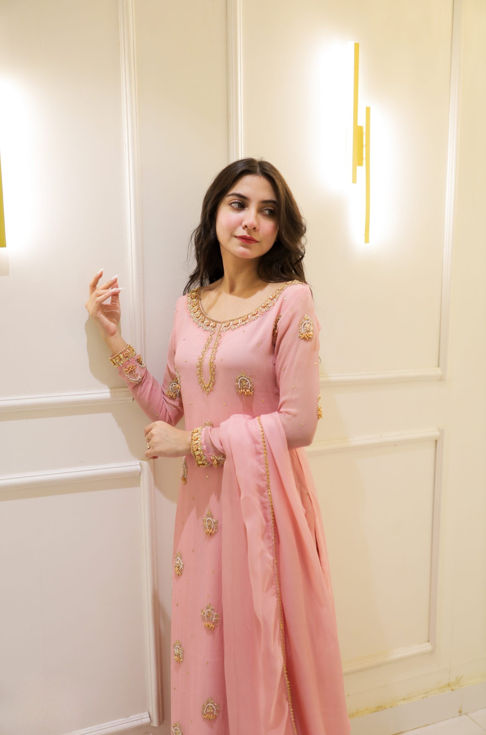 Woman in a pink traditional outfit with gold details against a white wall.