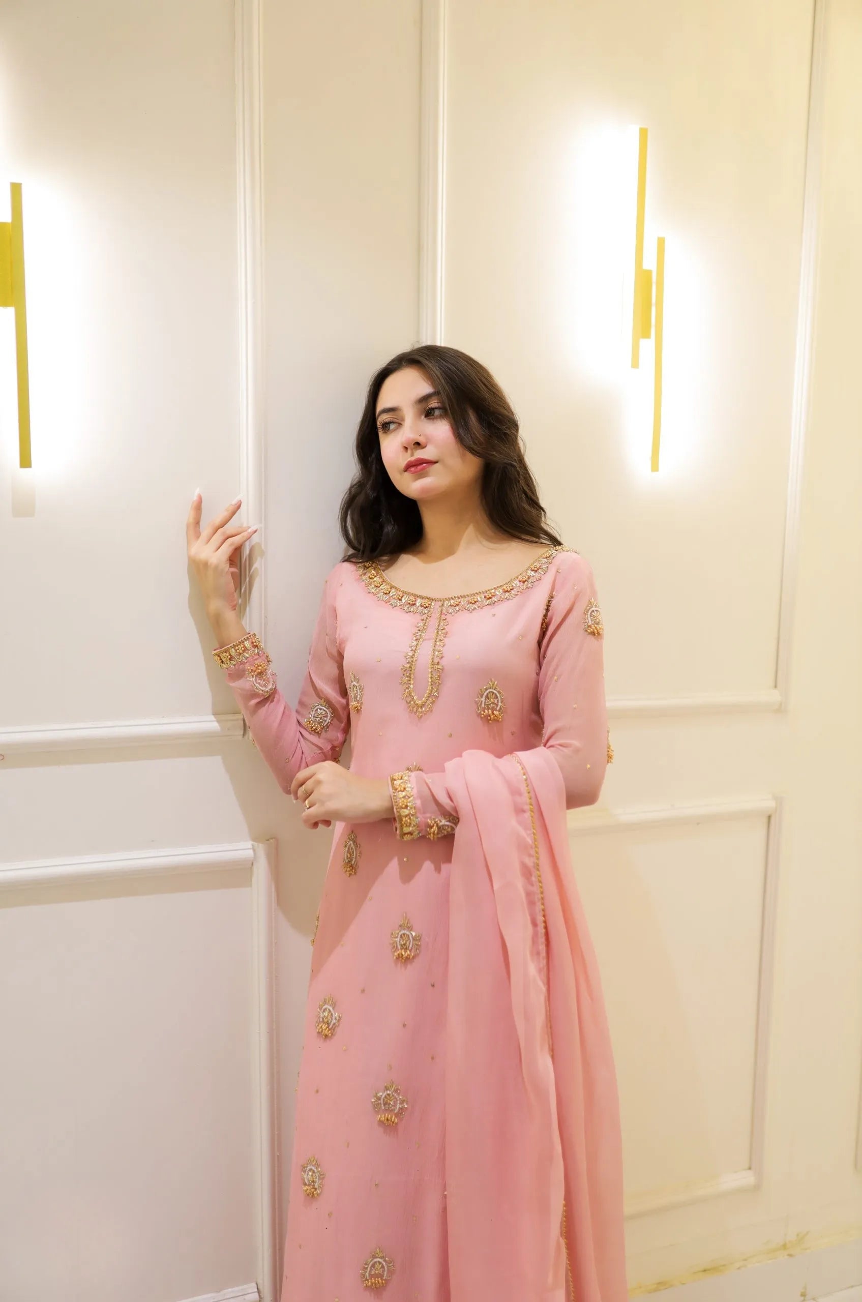 Woman in a pink traditional outfit with gold embroidery standing against a light-colored wall.