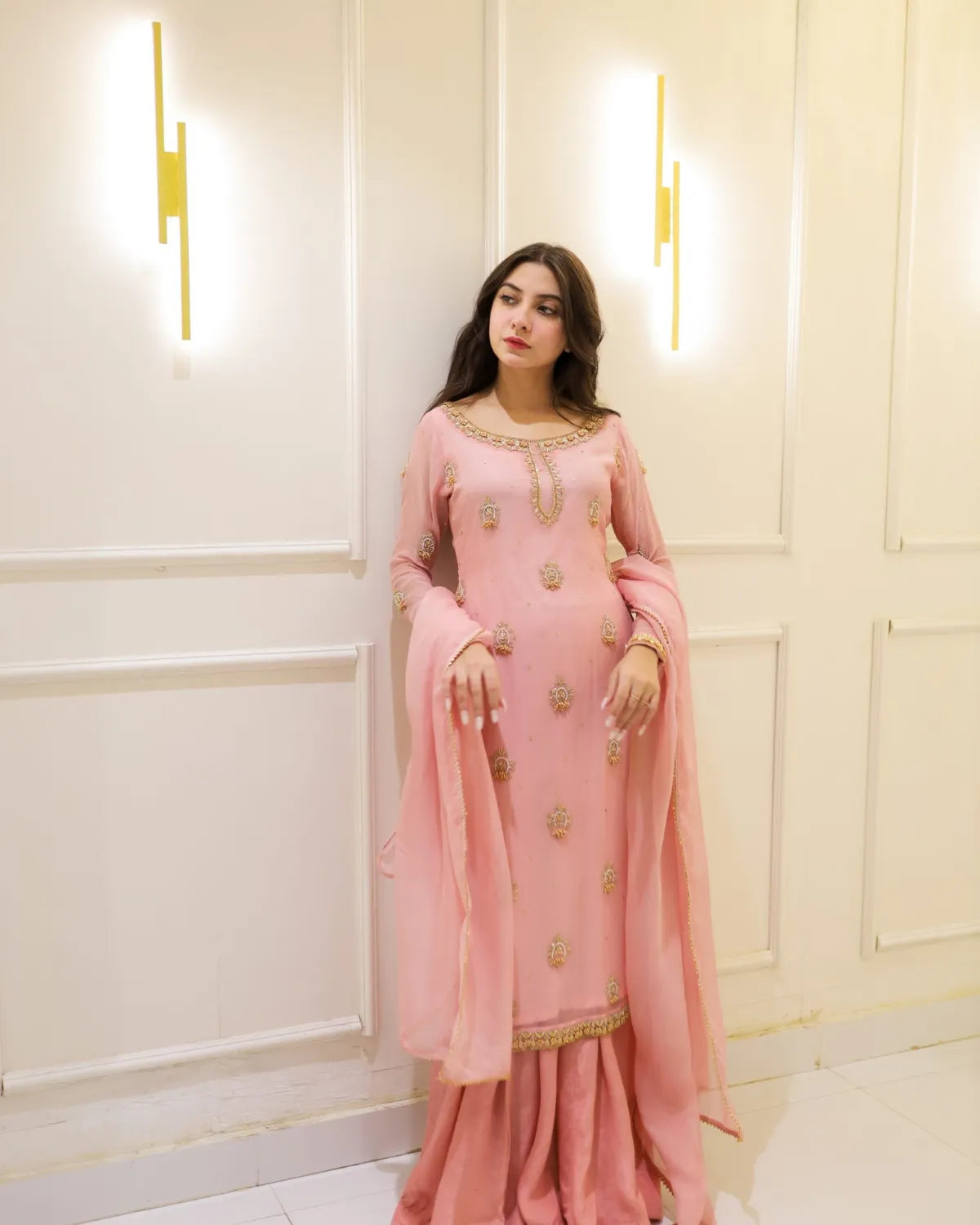 Woman in a pink traditional outfit with gold details standing against a light-colored wall.