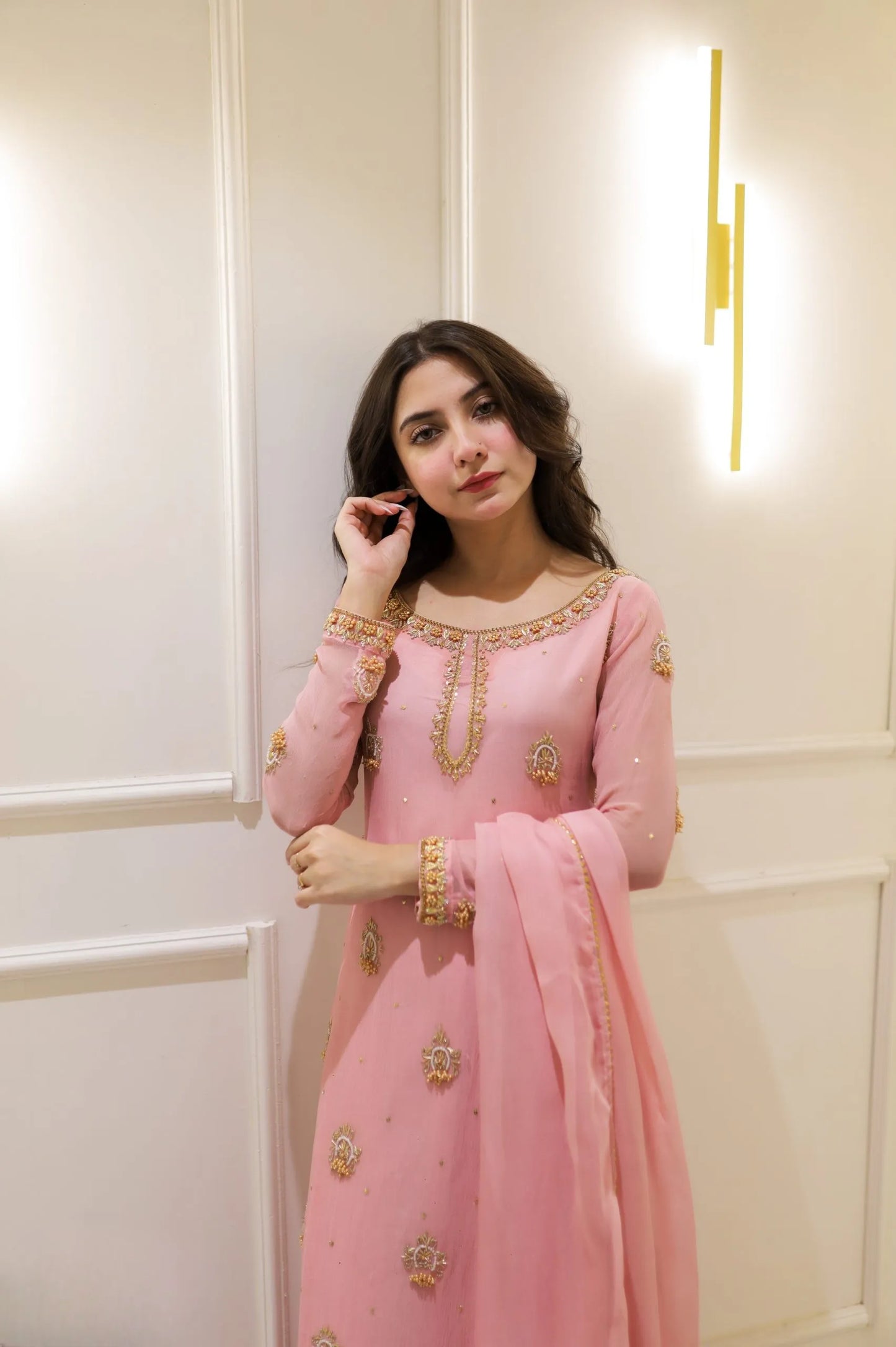 Woman in a pink traditional outfit with gold embroidery against a white wall.