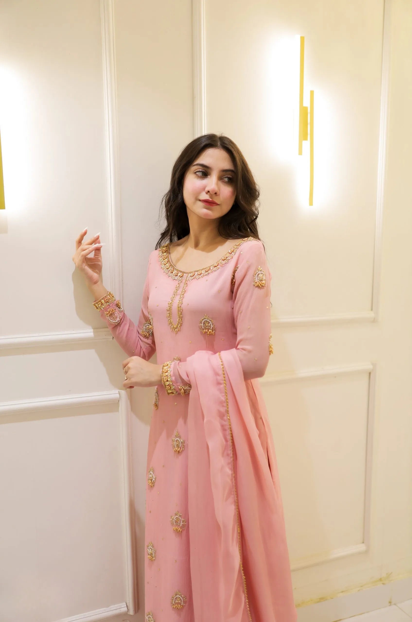 Woman in a pink traditional outfit with gold embroidery standing against a white wall.