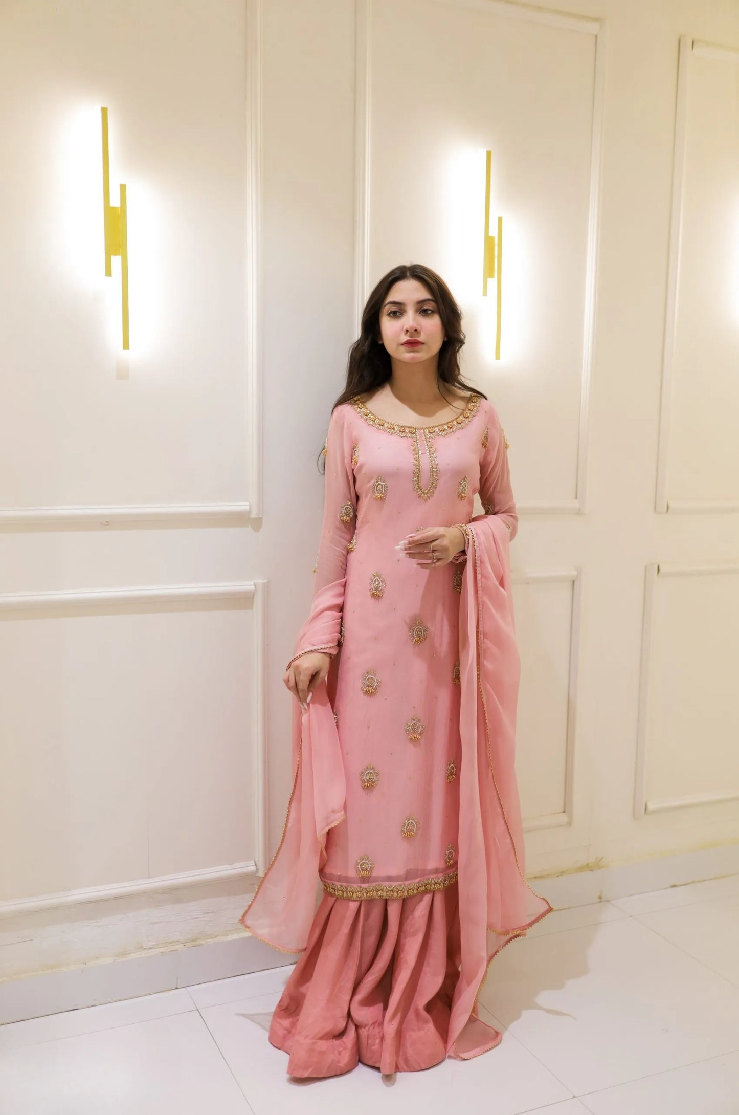 Woman in a pink traditional outfit standing against a white wall with decorative lights.