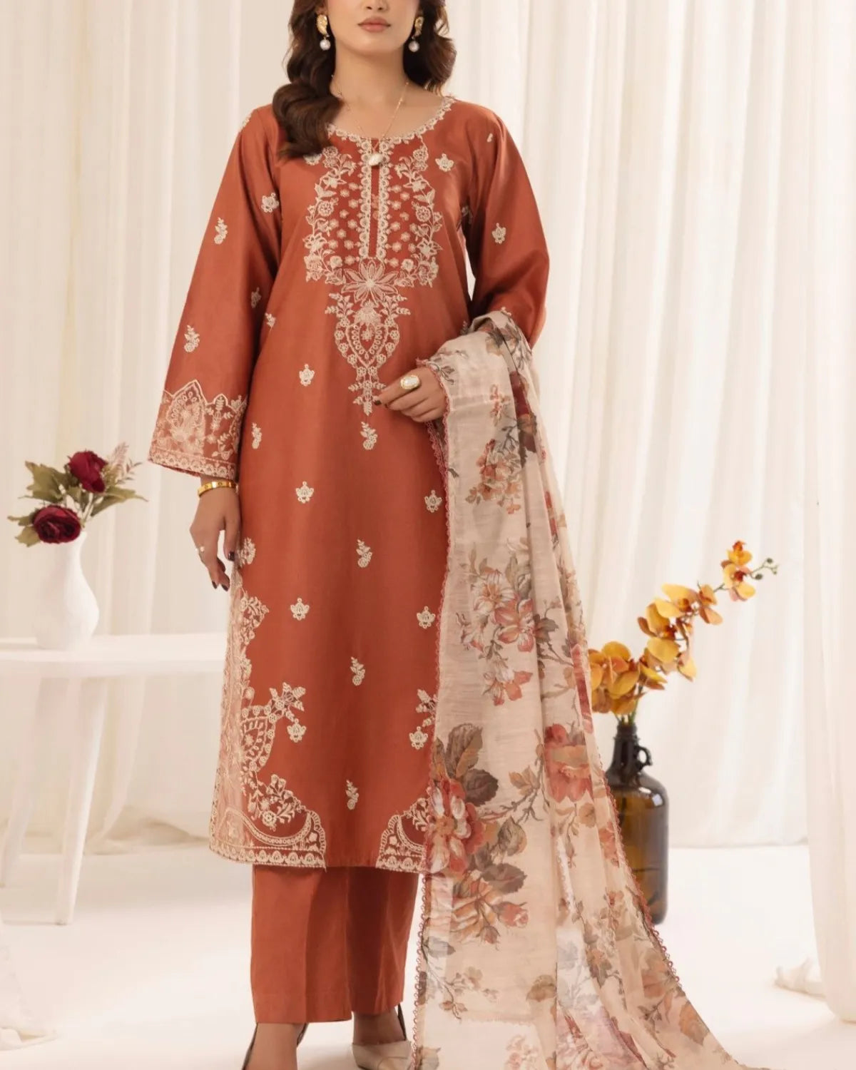 Woman wearing a rust-colored embroidered outfit with a floral dupatta against a white curtain background.