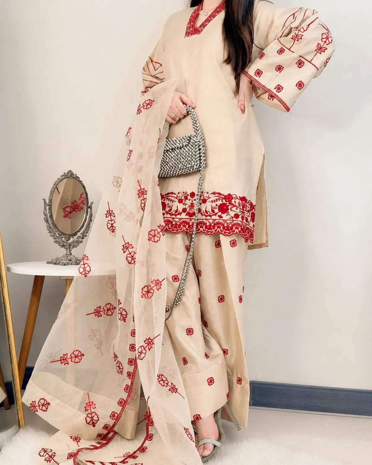 Woman wearing a beige outfit with red floral patterns, holding a handbag, against a plain background.