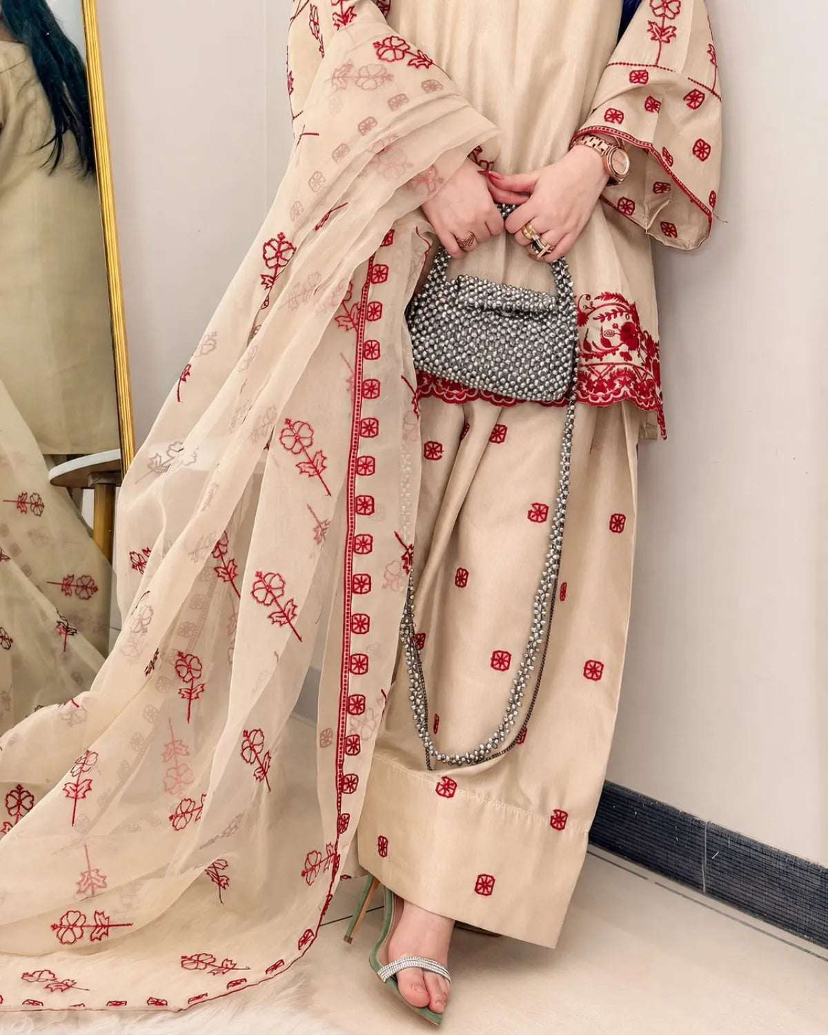 Person wearing a beige outfit with red patterns, holding a small handbag, standing in front of a mirror.