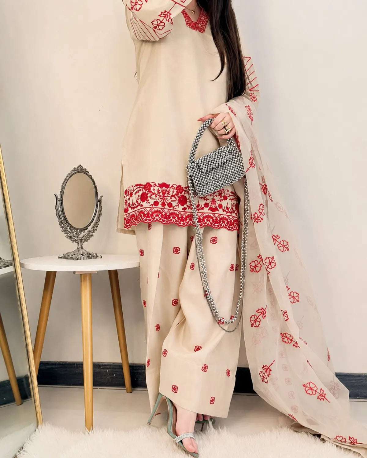 Woman in a cream outfit with red floral patterns holding a gray handbag in a minimalistic room.