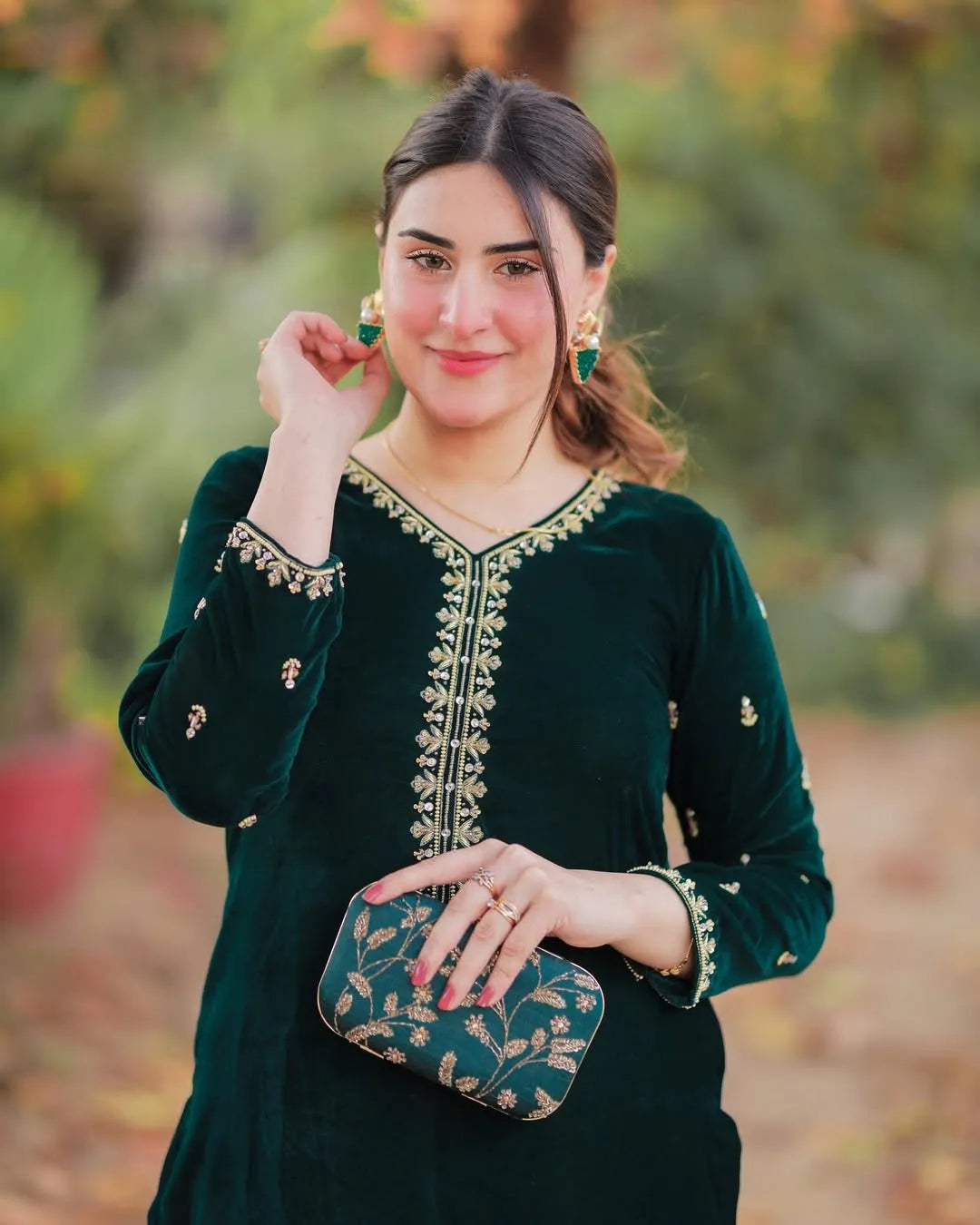 Woman in a green embroidered outfit holding a matching clutch against a blurred natural background