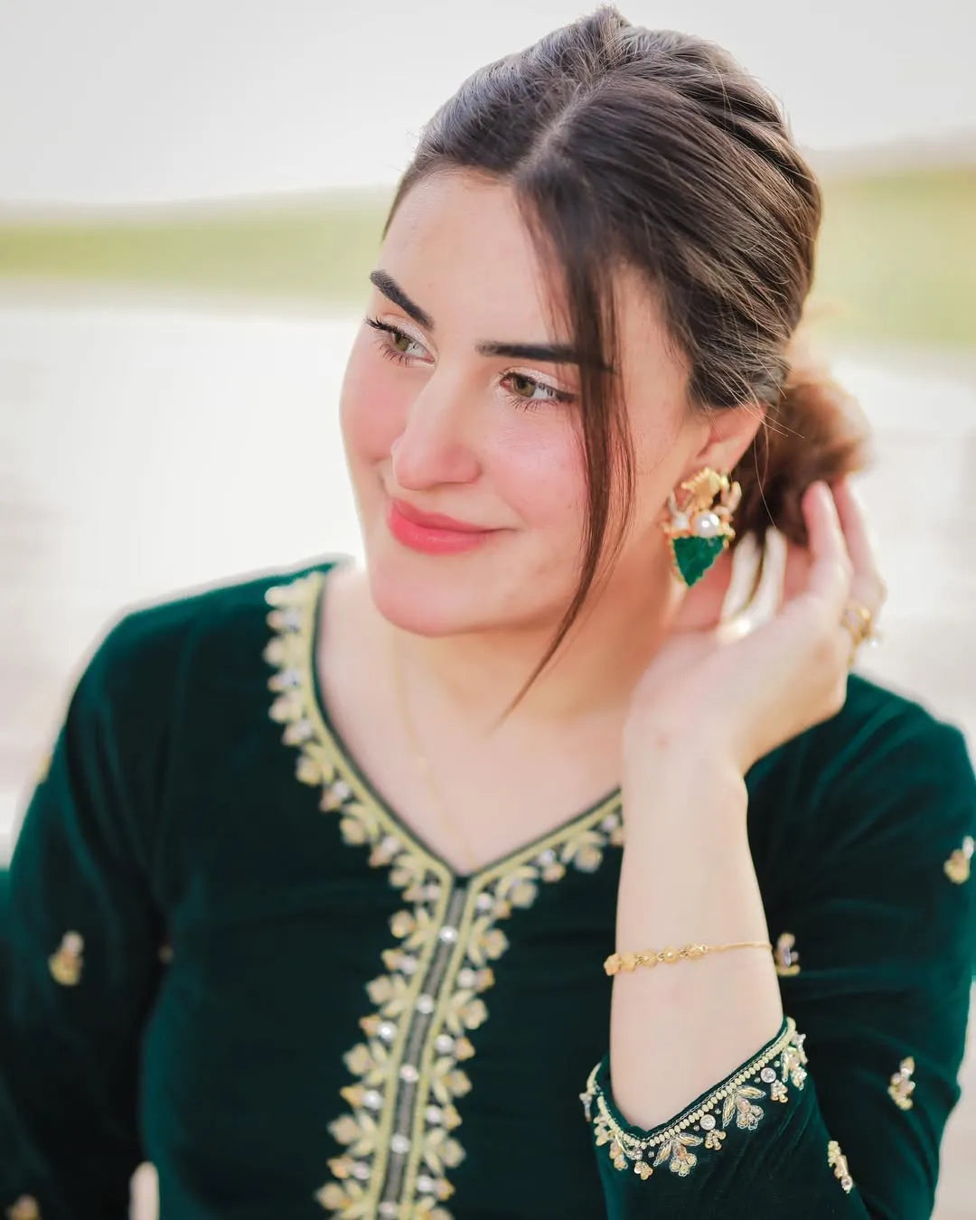 Woman wearing a green embroidered dress with gold jewelry, sitting outdoors.