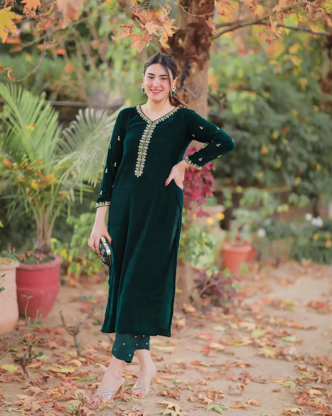 Woman in a green traditional outfit standing in a garden with trees and plants in the background.