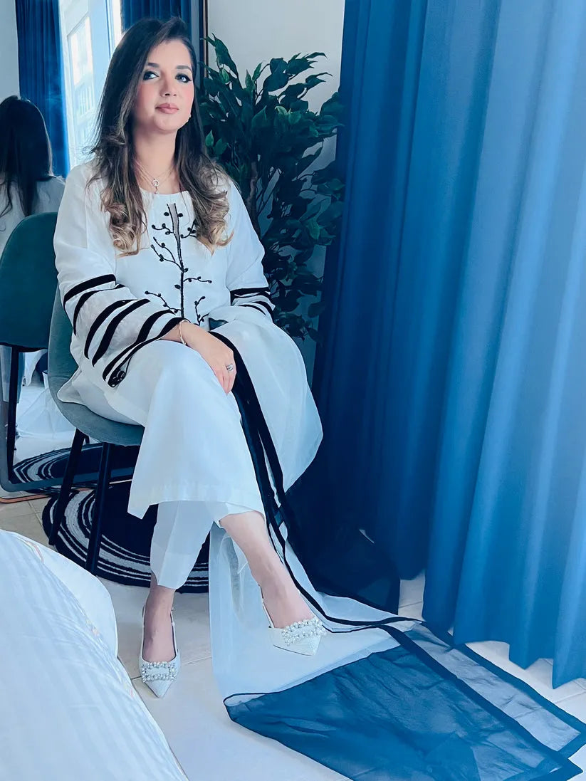 Woman in a white outfit with black accents sitting in a room with blue curtains and plants.