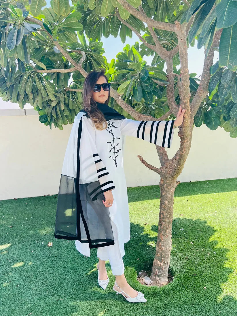 Woman in a white outfit with black accents standing next to a tree on grass.