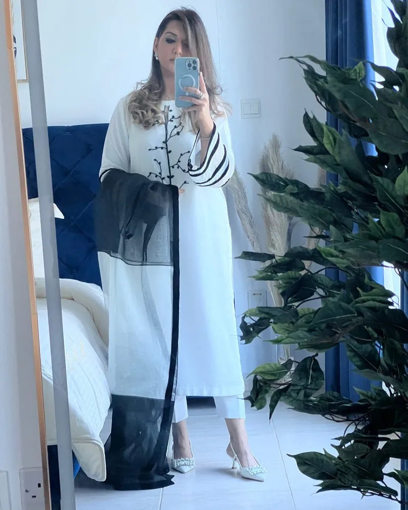Woman taking a mirror selfie wearing a long white dress with black accents in a room with plants.